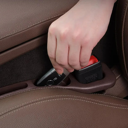 Hand placing a car key fob into the gap filler's storage slot beside the seatbelt buckle in a leather seat
