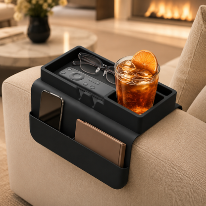 Black silicone couch cup holder on beige sofa armrest by a fireplace, holding an iced drink with orange slice, glasses, remote, phone and notebook — sleek living room organizer.