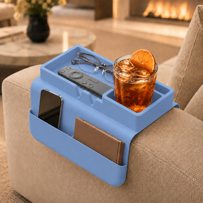 Light blue silicone couch cup holder on beige sofa armrest by a fireplace, holding an iced drink with orange slice, glasses, remote, phone and notebook — colourful living room sofa organizer.