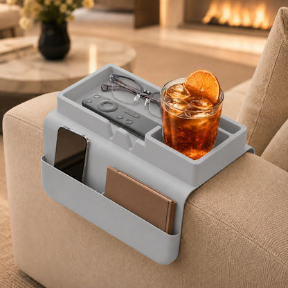 Grey silicone couch cup holder on beige sofa armrest by a fireplace, holding an iced drink with orange slice, glasses, remote, phone and notebook — modern living room sofa organizer.